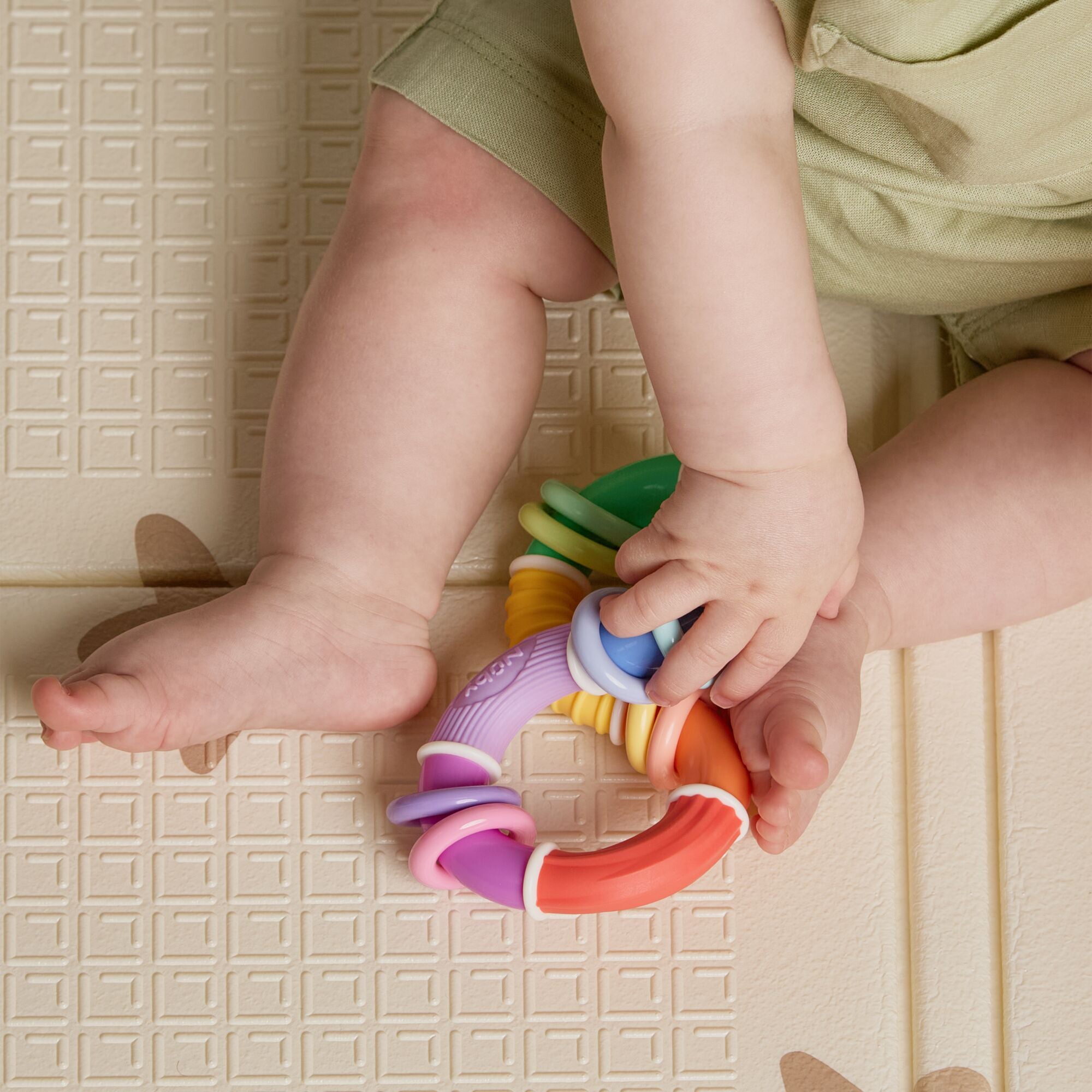 A baby in light green clothing sits on a textured mat with a simple square pattern, holding the Twista Rattle Teether Toy | Colorful Rings in both hands. Their legs are visible.