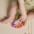 A baby in light green clothing sits on a textured mat with a simple square pattern, holding the Twista Rattle Teether Toy | Colorful Rings in both hands. Their legs are visible.