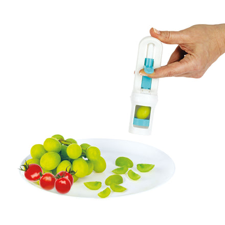 A hand uses the Nuby Grape Cutter | Pink to slice green grapes over a plate with whole and sliced grapes plus cherry tomatoes—ideal for preparing baby food and reducing choking risk.