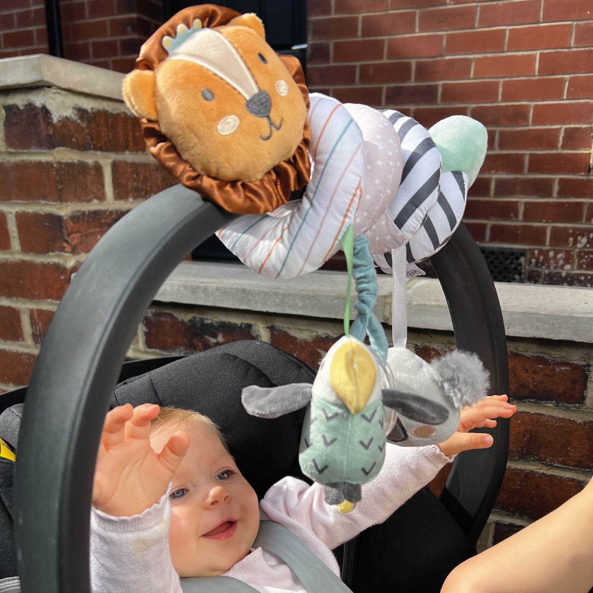 A smiling baby in a car seat reaches up to play with the Animal Adventures Spiral Activity Toy | Safari, which features plush lion and bird friends hanging from the handle, against a brick wall backdrop.