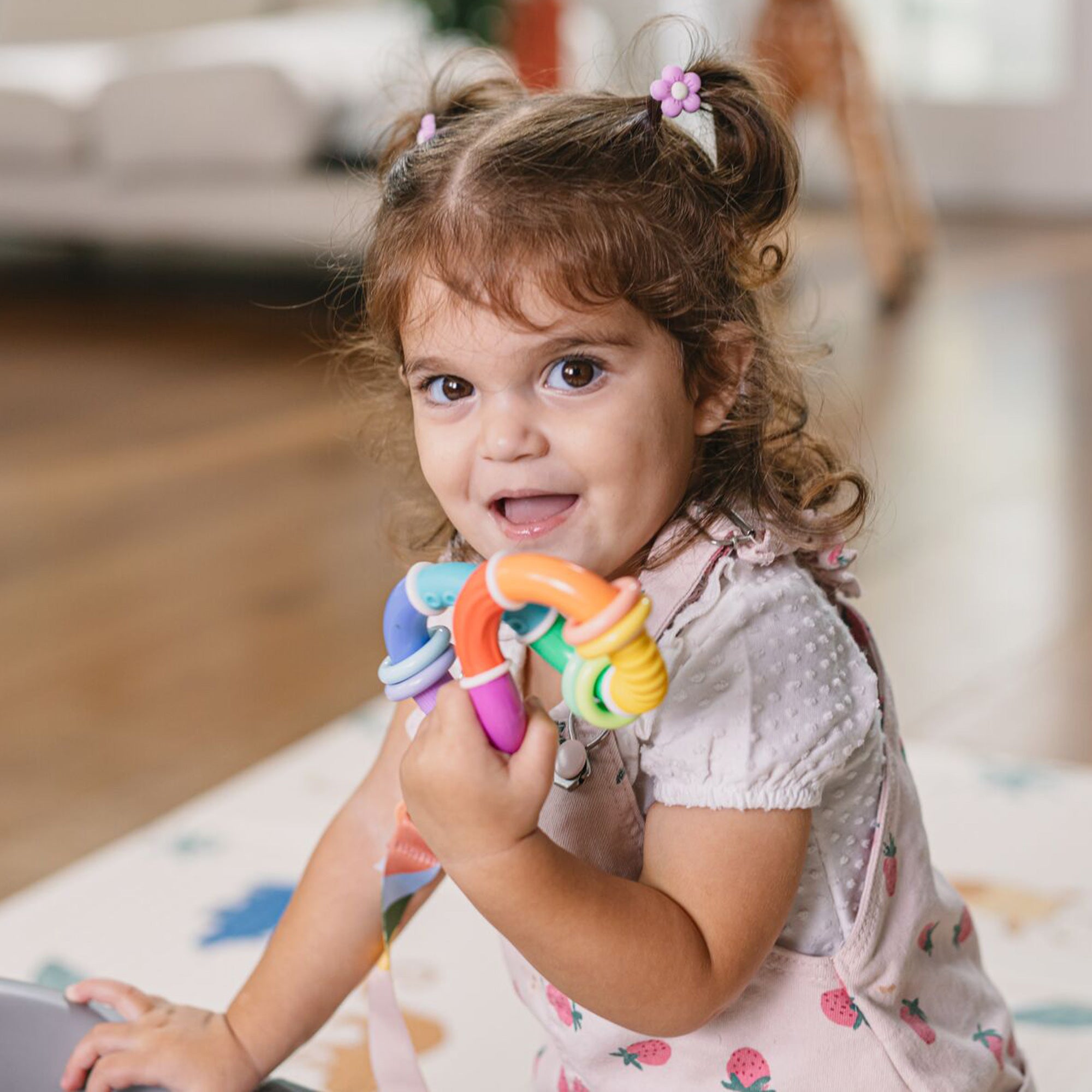 A young child with brown hair in pigtails, wearing a strawberry-print dress, smiles while holding the Twista Rattle Teether Toy | Colorful Rings in a bright, cozy room.