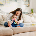 A woman sits on a beige couch with a baby in her lap. The baby holds the Twista Rattle Teether Toy | Colorful Rings, enjoying soothing teething relief and sensory play amid soft pillows and a plant for a cozy home vibe.
