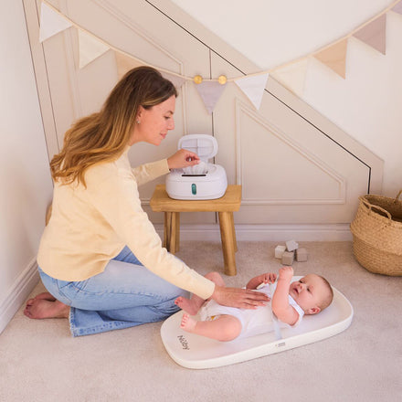 A woman changes her baby’s diaper on a Changing Pad with a non-slip base, placed on a carpeted floor. Nearby is a wooden stool holding wipes, while decorative bunting adorns the wall in the background.