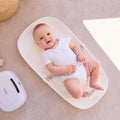A baby in a white onesie lies on the Changing Pad, secured with a gray safety harness, smiling up. A white device is on the floor nearby in the well-lit, cozy scene.