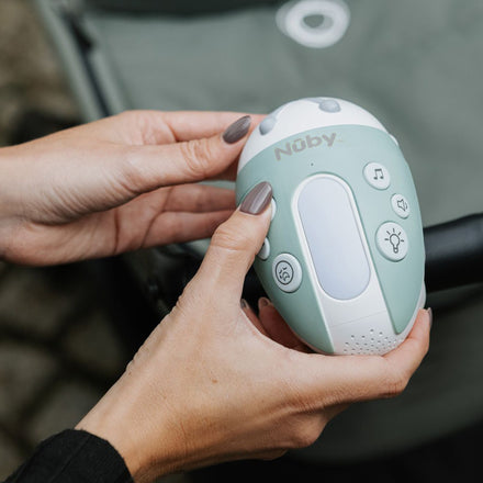 A person holds a mint green and white Nuby Jitterbug Rocker with music, light, and settings buttons, positioned above a stroller or baby gear in the background.