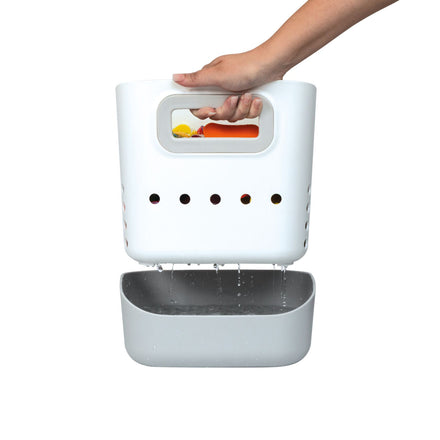 A hand holds the Nuby Tub Toy Caddy, a white plastic organizer with drainage holes, letting water flow into a gray base. The caddy is filled with colorful bath toys.