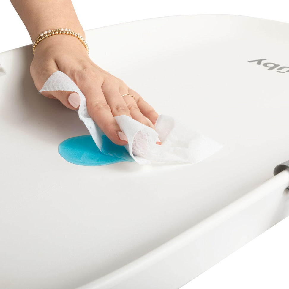 A hand wearing a beaded bracelet and a ring uses a white paper towel to wipe up a blue spill on the smooth, non-slip base of the Changing Pad.