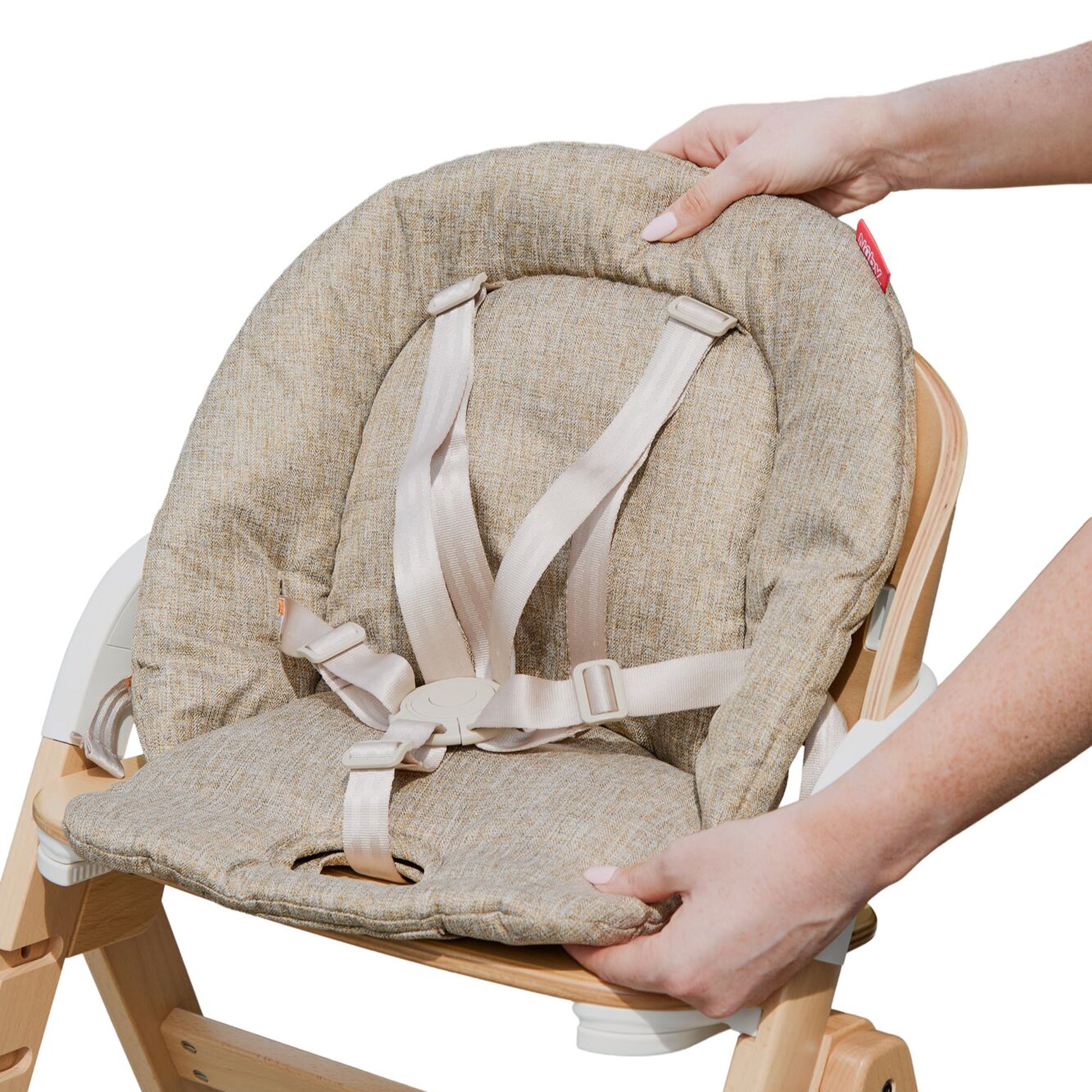 A person adjusts a beige cushioned Grow with Me Foldable High Chair with safety straps, attached to a wooden frame.