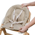 A person adjusts a beige cushioned Grow with Me Foldable High Chair with safety straps, attached to a wooden frame.