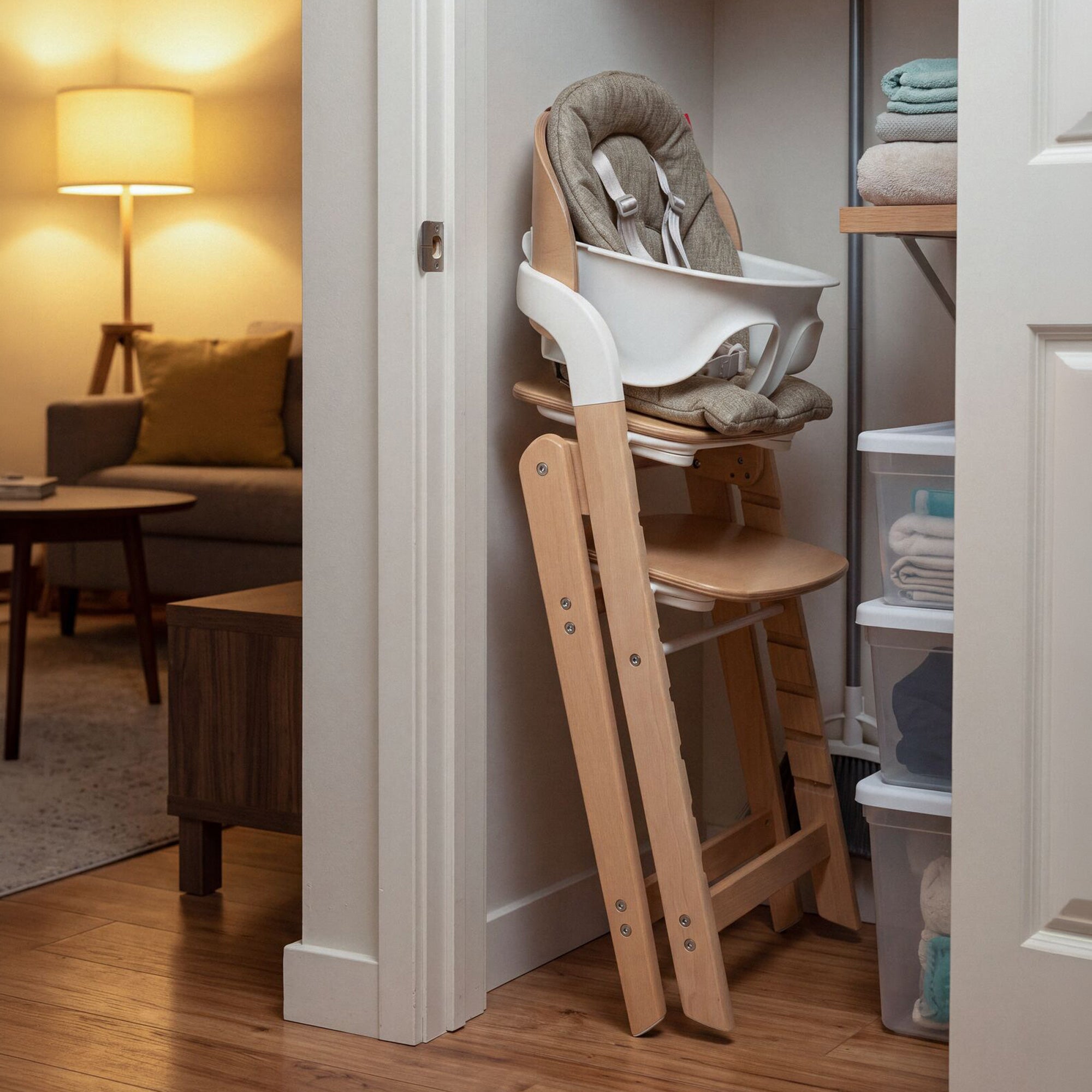 A Grow with Me Foldable High Chair, featuring a cushioned seat and tray, is folded and stored in a closet next to shelves with towels, while a lit living room can be seen in the background.