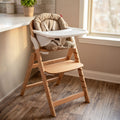 The Grow with Me Foldable High Chair, featuring a padded seat and a white tray, sits beside a kitchen counter near a window on a wooden floor in a sunlit room.