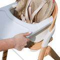 A hand adjusts the white tray of the Grow with Me Foldable High Chair, which features a wooden frame, beige fabric seat, and safety straps.