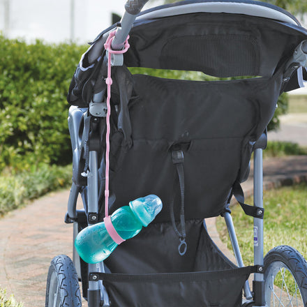 A blue baby bottle is secured to a black stroller with a pink strap from the Nuby Pacifier & Bottle Bungee Set (4 Pack); the stroller is outdoors on a paved path surrounded by greenery.