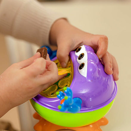 A child plays with the Nuby iMonster Snack Keeper, a spill-proof snack container featuring colorful purple and green pop-it bubbles, googly eyes, and a blue animal figure on its side while holding a small brown object.
