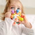 A young child in a white shirt holds and chews on the Twist Ball Teether, a colorful sensory toy with various textured shapes, gazing upward with blue eyes.