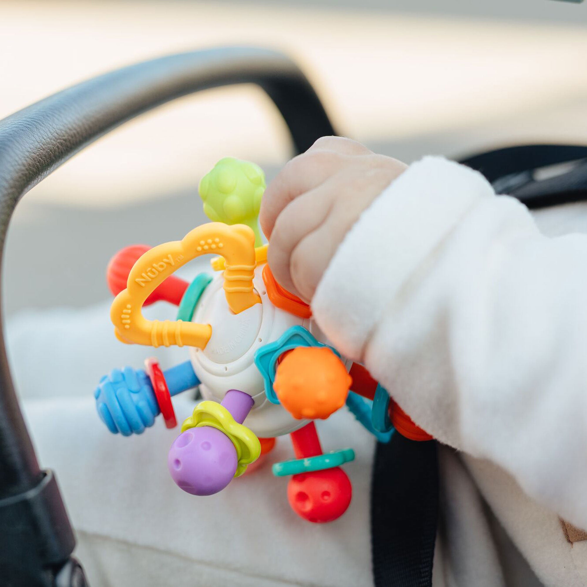 A baby, dressed in white and sitting in a stroller, holds the Twist Ball Teether—a colorful and multi-textured toy.