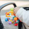 A baby, dressed in white and sitting in a stroller, holds the Twist Ball Teether—a colorful and multi-textured toy.