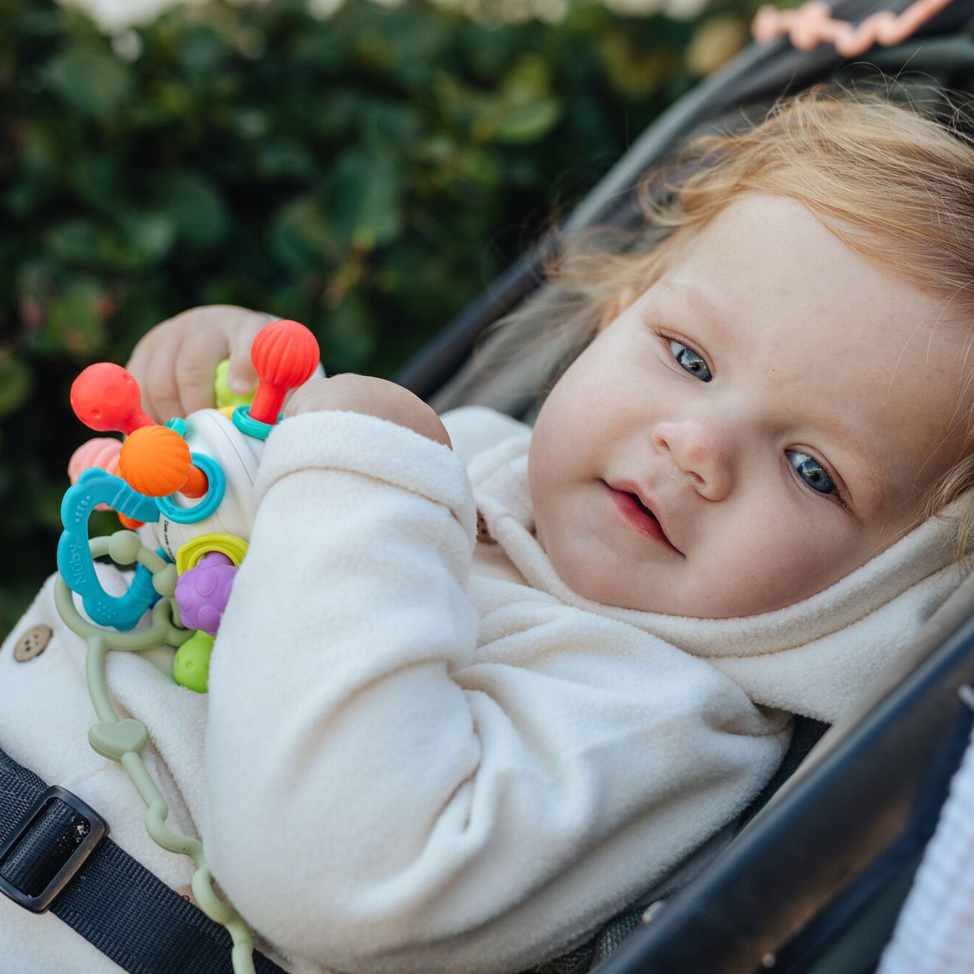 A baby with light hair and blue eyes, dressed in white, sits in a stroller and plays with the colorful Twist Ball Teether. Blurred green foliage is visible in the background.
