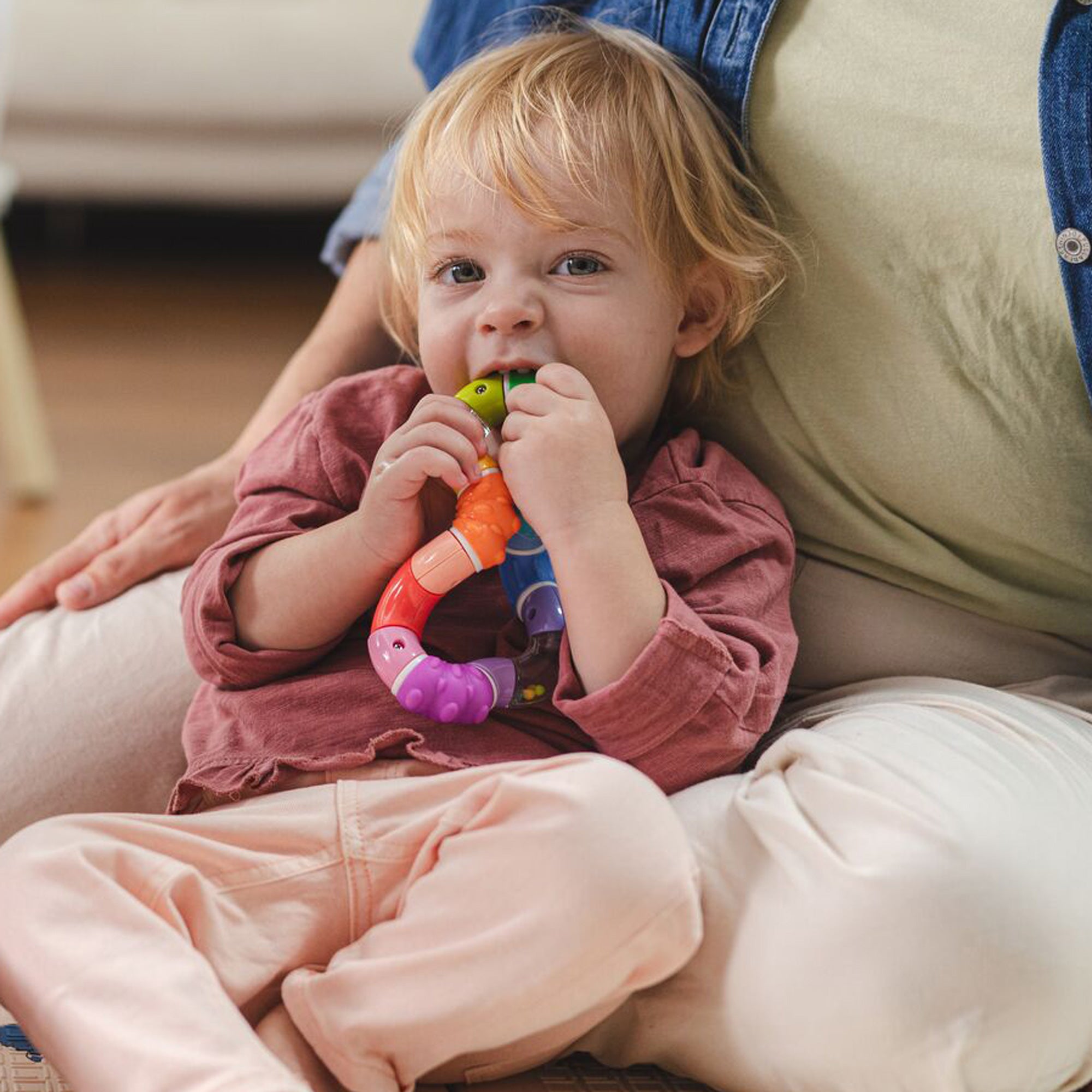A blonde-haired toddler in a pinkish shirt and light pants sits on an adult’s lap, curiously looking at the camera while chewing on and enjoying the colorful Twista Rattle Teether Toy | Rattle Balls.