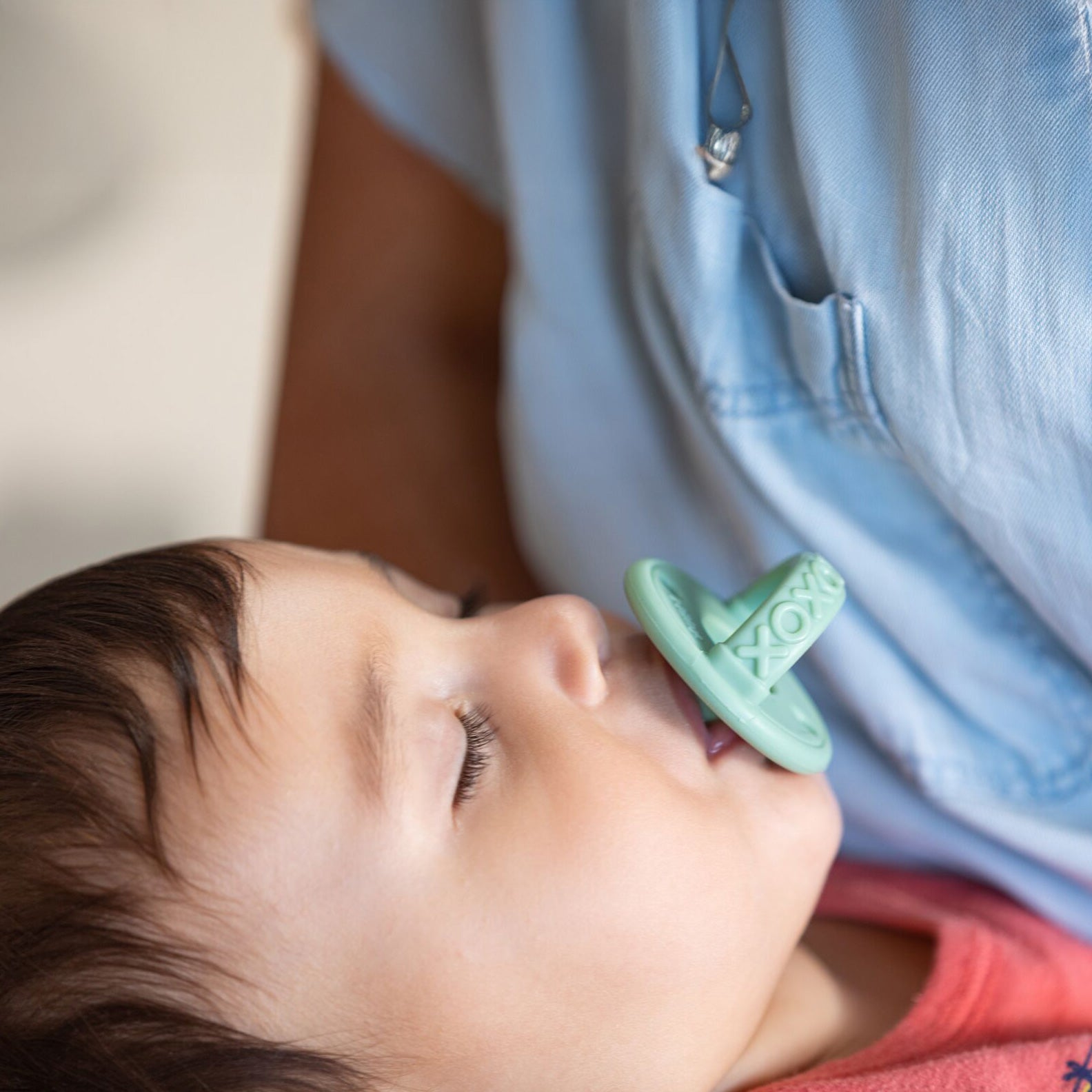 A baby with closed eyes sucks on a green Nuby Sili Soother Pacifier (2 Pack) while resting against an adult in a light blue shirt.
