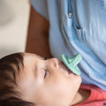 A baby with closed eyes sucks on a green Nuby Sili Soother Pacifier (2 Pack) while resting against an adult in a light blue shirt.