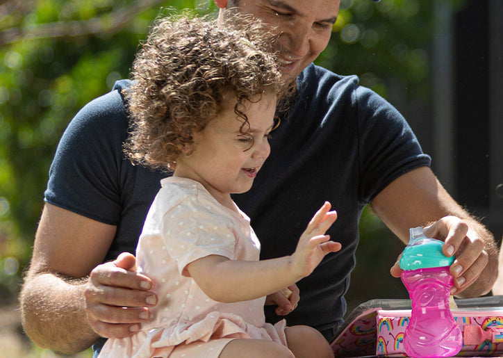 A father holding a pink Nuby sippy cup for his toddler daughter as she reaches for it.