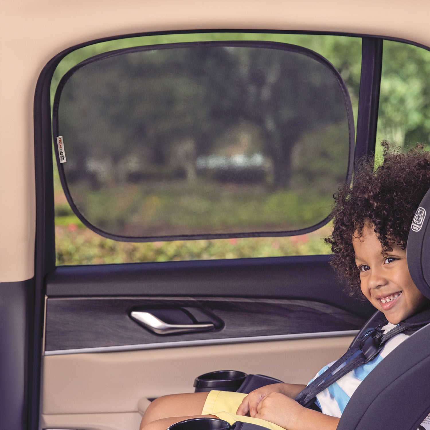 A smiling child sits in a car seat by the window, which is covered with a Safe in the Seat Pop Open Cling Sunshade (2 Pack) offering UV protection. Trees and greenery are visible outside.