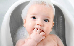 Baby with fingers in mouth, soap suds on head, sitting in a grey Nuby Non-Slip Bathing Cradle.