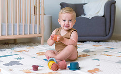Sitting toddler playing with stacking toys on a safari-print Nuby Reversible Floor Mat.