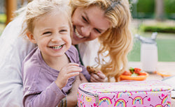 Mother hugging a smiling girl who is eating a cookie. A rainbow-patterned Nuby Insulated Bento Box Lunch Box is on the table.