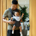 A smiling man stands indoors, carrying a cheerful baby in the Nuby Classic Baby Carrier | Black, made with breathable mesh. The baby, in a patterned gray bib with raised arms, delights as the man looks down—ideal from newborn to toddler.