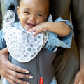 A smiling baby wearing a blue outfit and dotted white bib is held by an adult in the Nuby Classic Baby Carrier | Grey, looking comfortable and content from newborn to toddler stage.