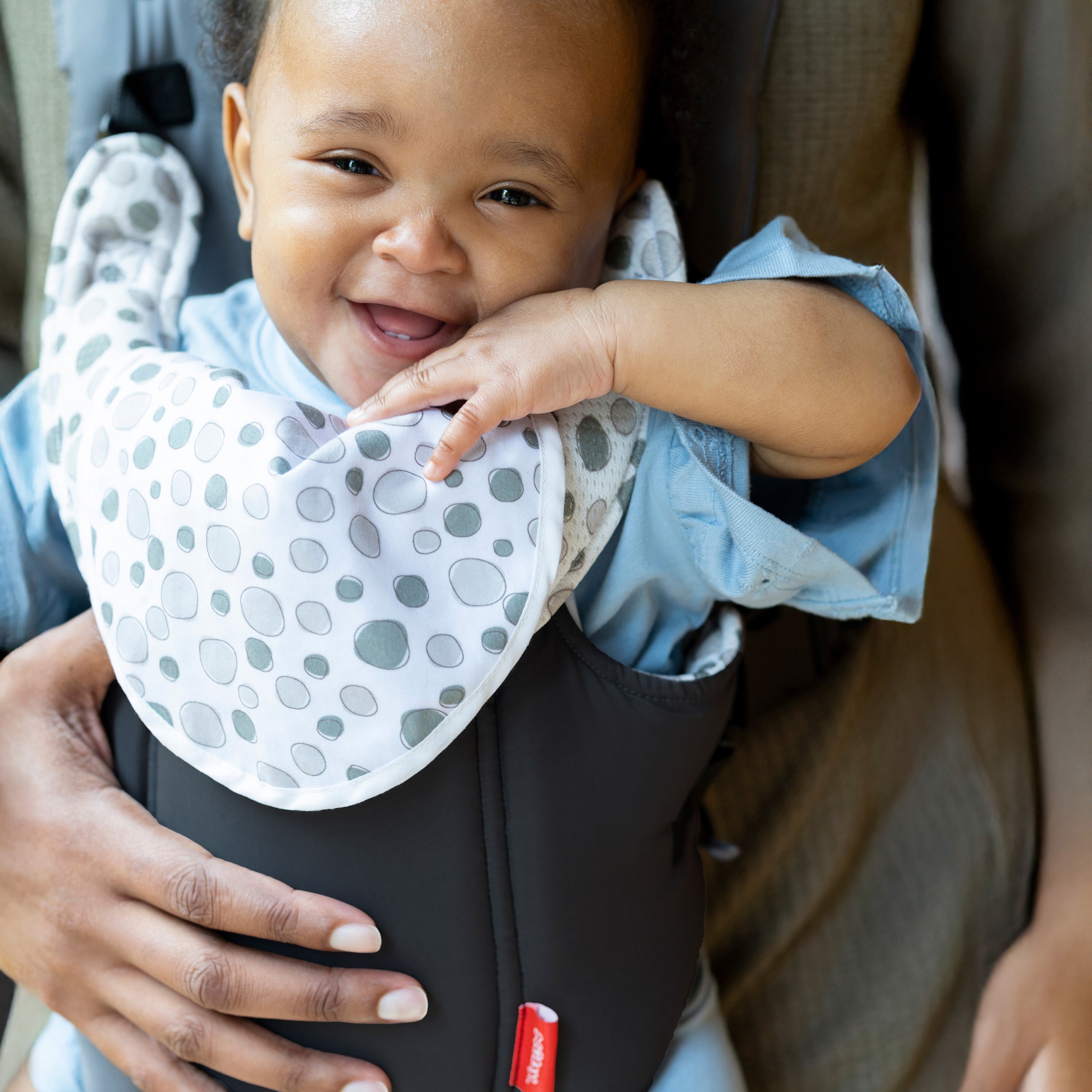 A smiling baby in a blue outfit and white bib is held securely in the Nuby Classic Baby Carrier | Black, featuring breathable mesh—perfect for newborns to toddlers.