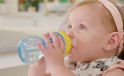 Baby girl with pink headband drinking water from a clear blue Nuby Mini Gripper Soft Spout Sippy Cup.