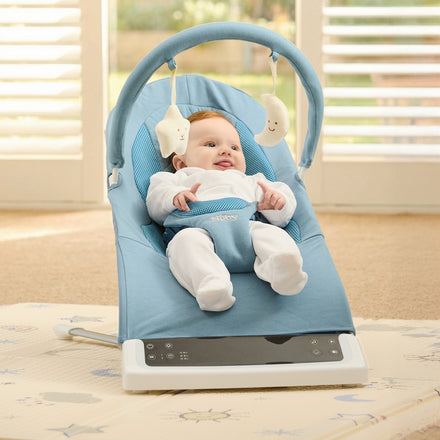 A smiling baby enjoys the bright, sunlit room while sitting in the Deluxe Baby Bouncer Seat | Blue, featuring an overhead toy arch. The floor is covered with a white mat decorated with small stars and animal designs.
