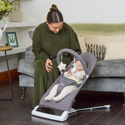 A woman in green sits on a gray sofa, watching a baby drink from a bottle while resting in the Deluxe Baby Bouncer Seat | Grey in a cozy living room.