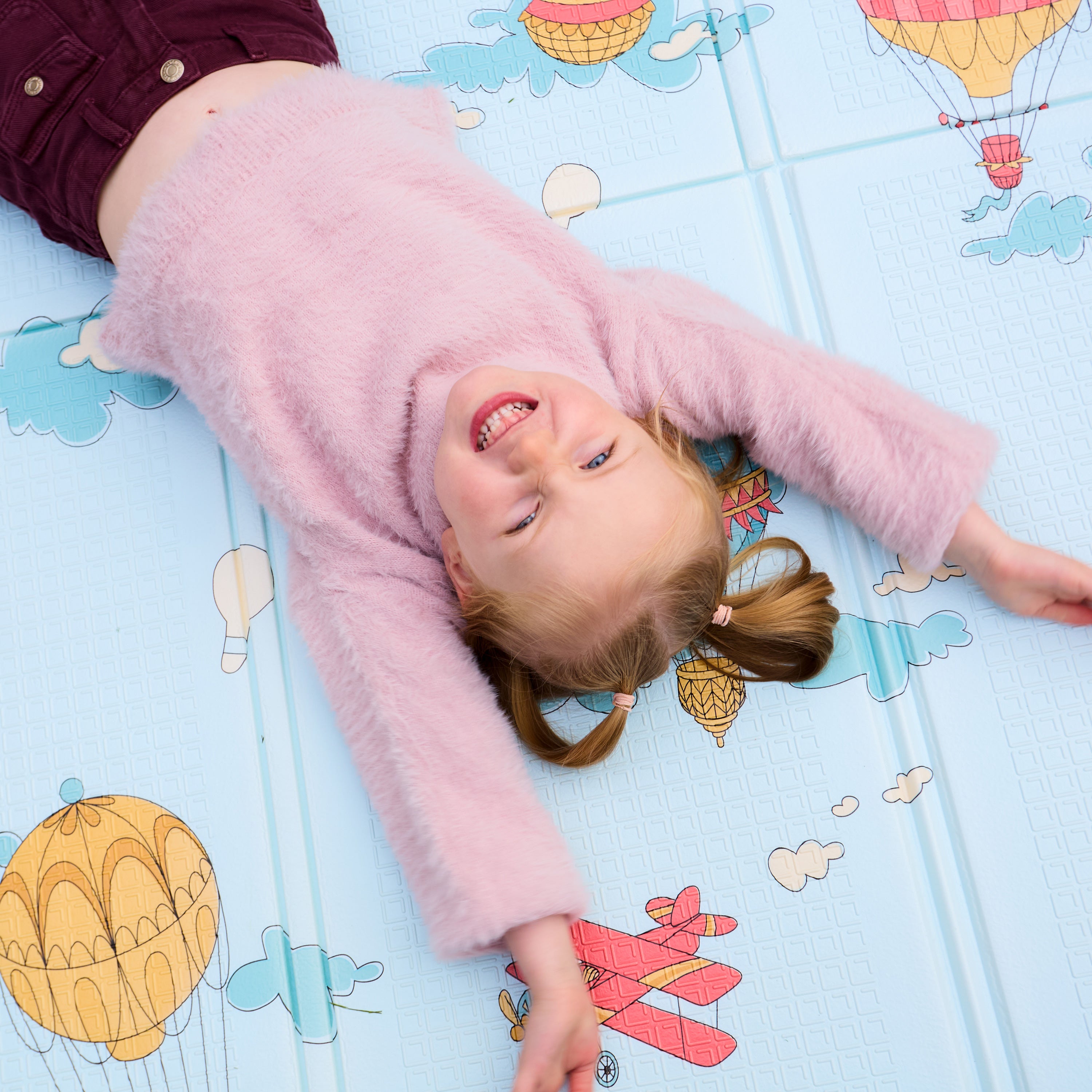 A young child with light brown pigtails, in a fuzzy pink sweater and maroon pants, smiles with arms outstretched while lying on the Reversible Floor Mat | Hot Air Balloons, which features colorful hot air balloons and clouds.