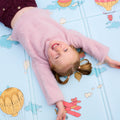 A young child with light brown pigtails, in a fuzzy pink sweater and maroon pants, smiles with arms outstretched while lying on the Reversible Floor Mat | Hot Air Balloons, which features colorful hot air balloons and clouds.