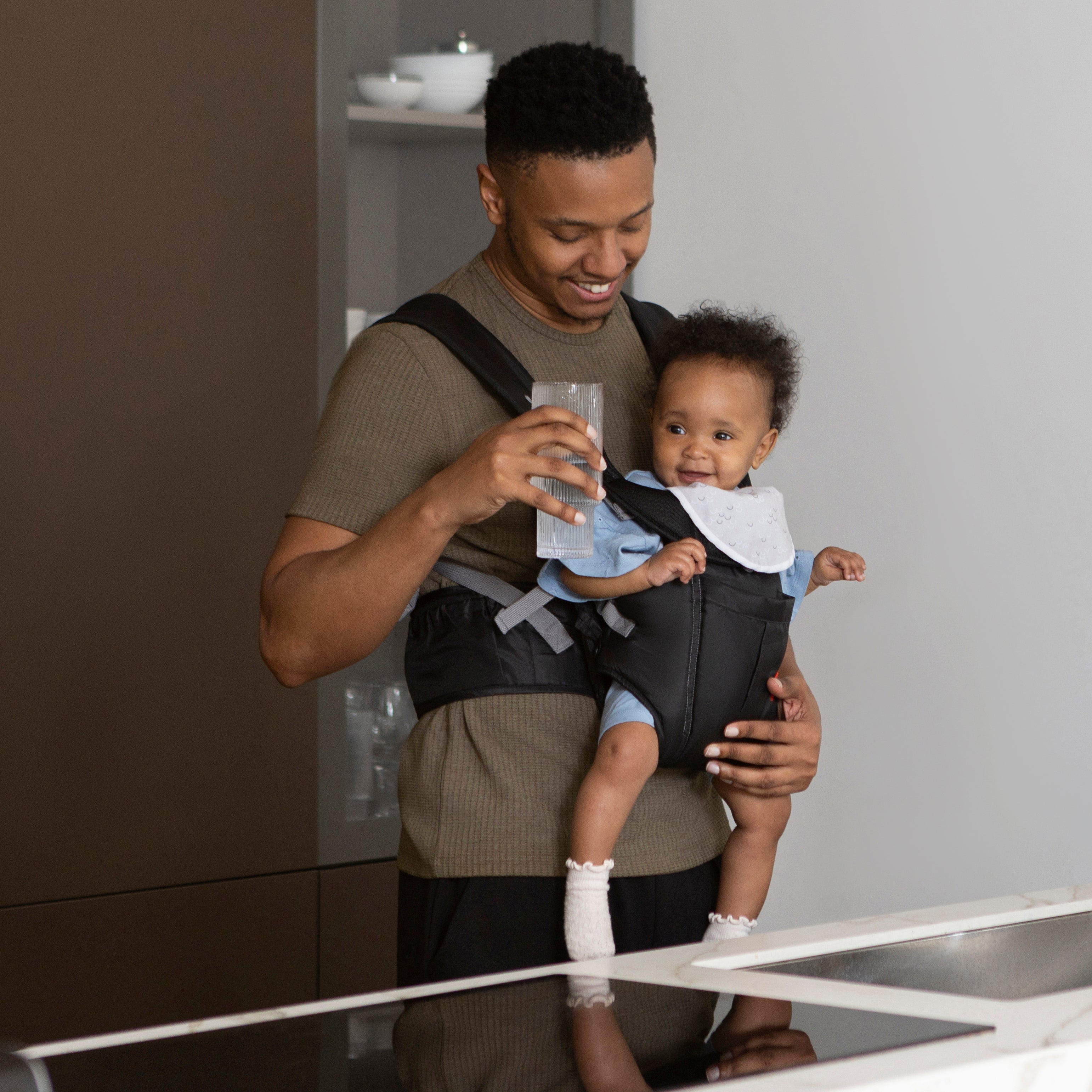 A smiling man in a modern kitchen holds a glass of water and carries a happy baby in the Deluxe Baby Carrier | Black, featuring adjustable straps.