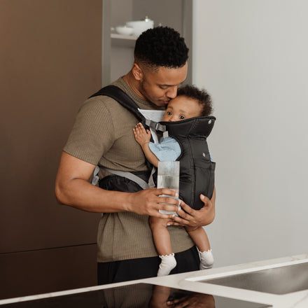 A man stands in a kitchen, holding a glass of water while carrying a baby in the Deluxe Baby Carrier | Black. The adjustable straps secure the baby comfortably against his chest, and both look relaxed and content.