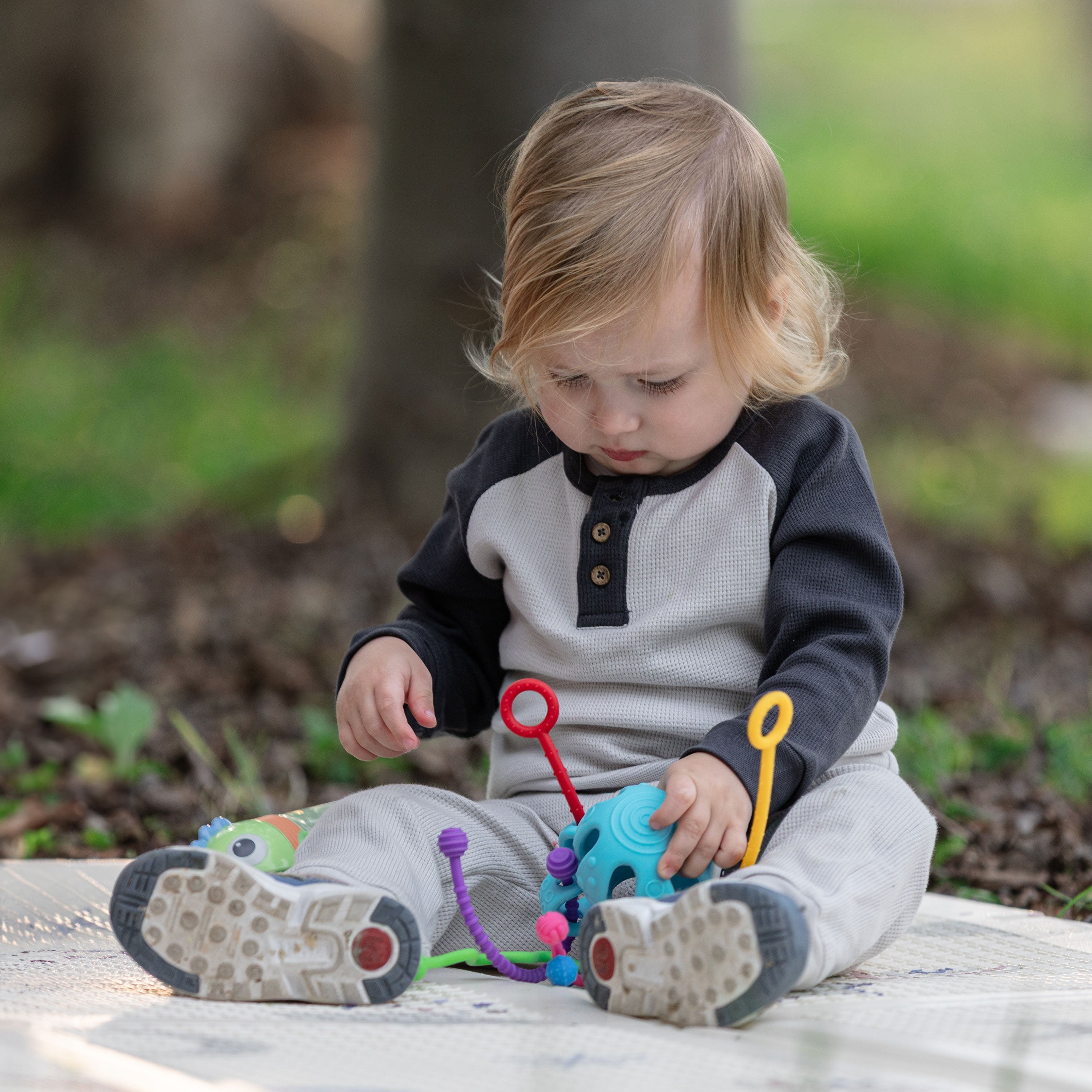 A young child with light brown hair sits on a blanket outdoors, playing with the Octopus Silicone Toy. Dressed in a gray and black long-sleeve outfit, the child supports tactile development and motor skills on the grass among trees.