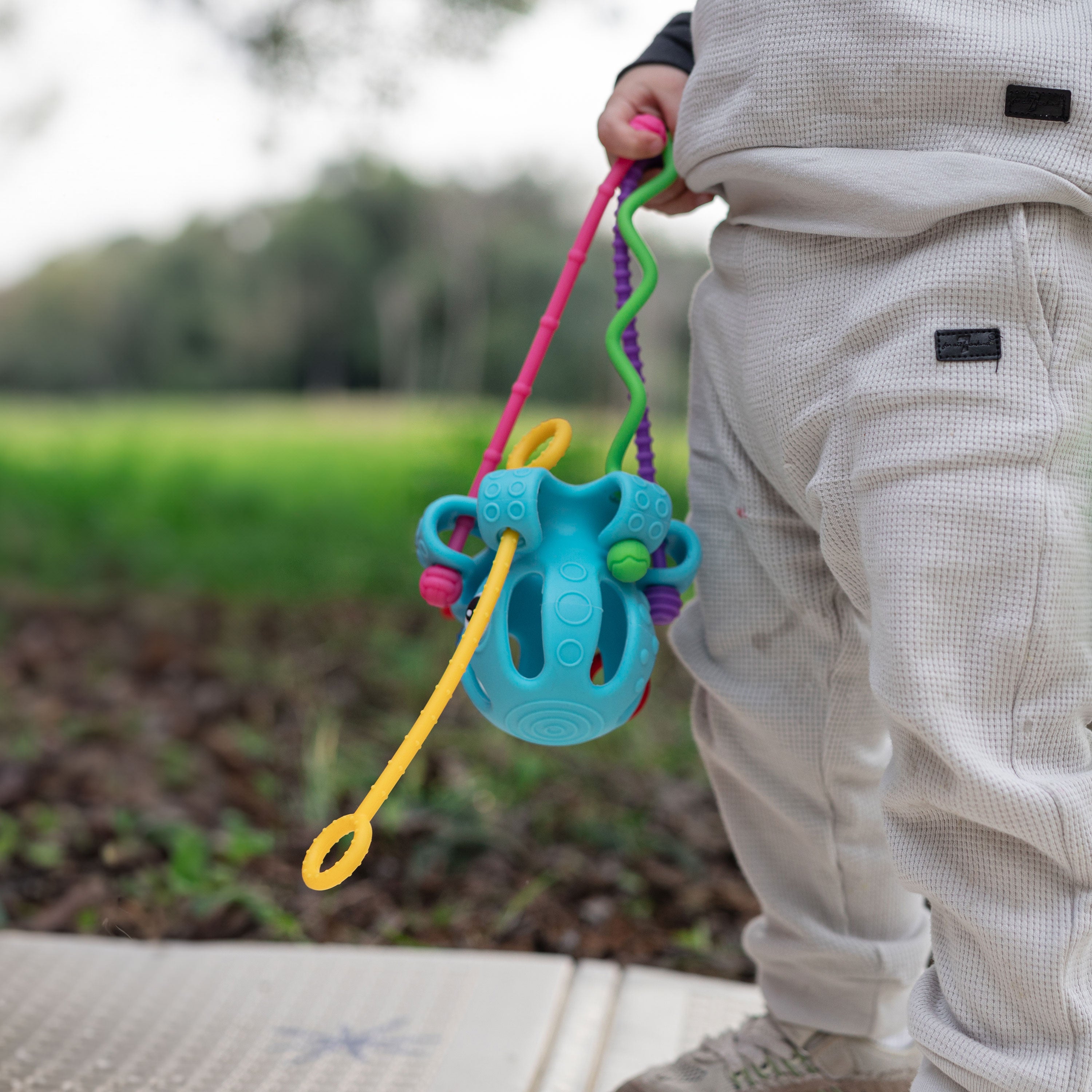 A young child in light-colored clothes stands outdoors holding the Octopus Silicone Toy, supporting motor skills and tactile development, with grass and trees softly blurred in the background.