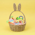 The Nuby US One & Done Bunny Basket Bundle features a woven basket with bunny ear handles, colorful toys including a Bunny Basket Teether and plush carrot, plus pink paper shreds, perfect for sensory play on a yellow background.