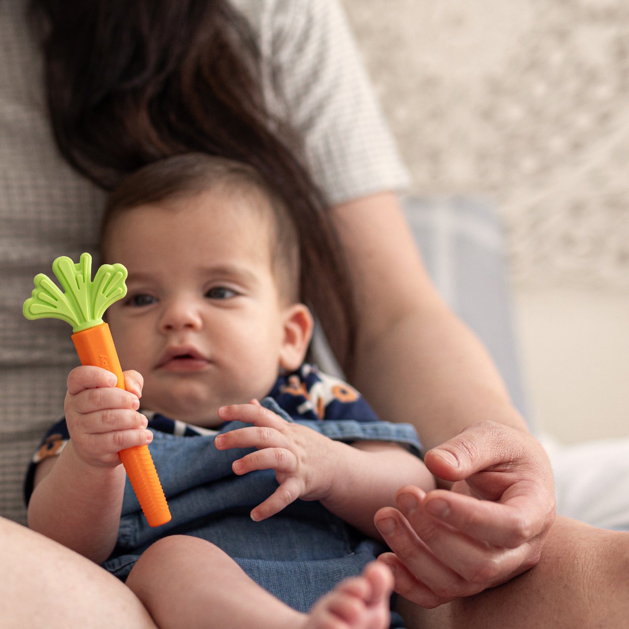 A baby sits on an adult’s lap, reaching for the green and orange carrot-shaped teether from the One & Done Bunny Basket Bundle by Nuby US, while the adult gently supports during this sweet moment of sensory play.