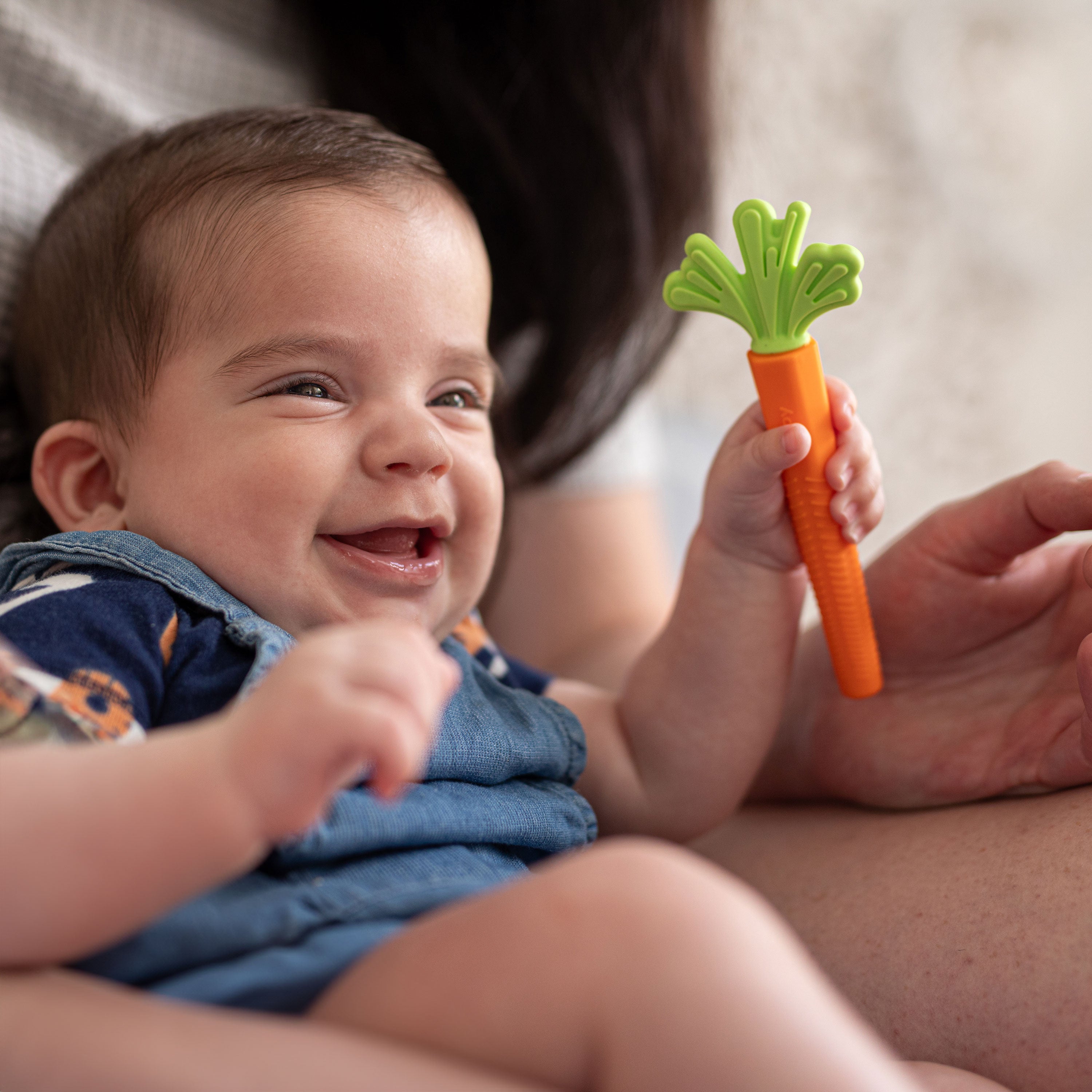 A happy baby in blue overalls sits on an adult's lap, holding the Silicone Carrot Tube Teether. The colorful teether provides gentle relief for little ones, making teething time easier and more playful.