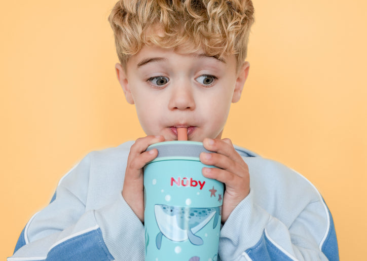 A young boy with curly blond hair in a light blue sweater drinks from the Nuby Thirsty Kids Flexstraw Stainless Steel Cup | Whales, looking surprised against a solid yellow background.