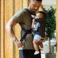 A man adjusts the strap of the Nuby Classic Baby Carrier | Black, featuring breathable mesh, while holding a baby from newborn to toddler age. They stand indoors near a wooden wall and a potted plant.