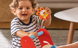 Toddler holding a Nuby Vibe-eez Vibrating Baby Teether (Lion) with one hand and pushing a Nuby Wooden Baby Walker with the other.