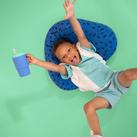 A smiling child in teal shorts and a white shirt lies on a blue cushion, arms outstretched and tongue out, holding the Silicone Tiny Tumbler in Periwinkle with its spill-proof straw, against a mint green background.