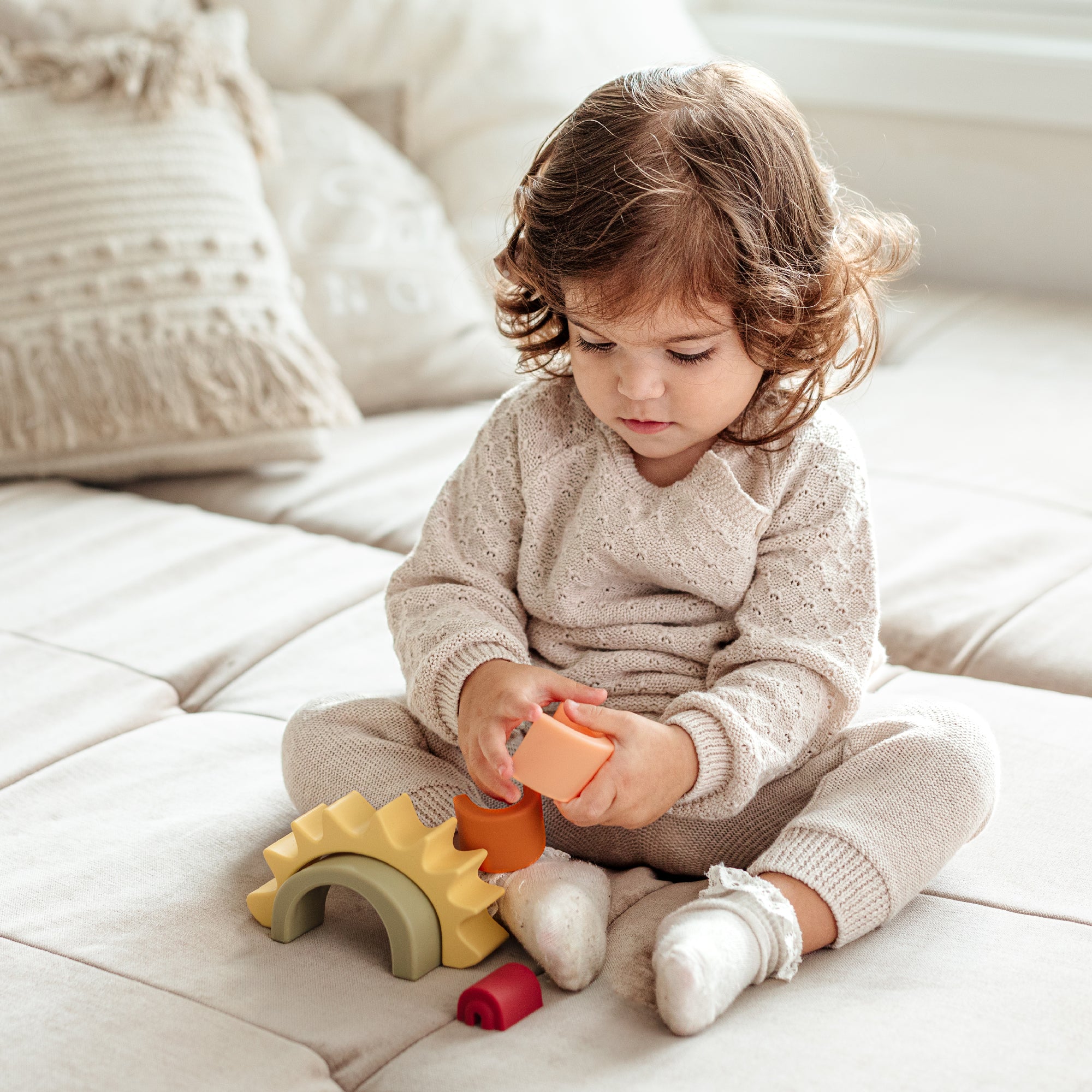 A toddler with curly hair plays with the Nuby US One & Done Bunny Basket Bundle, a sensory play set featuring colorful arches and cylinders, while sitting on a beige sofa with cozy cushions and an Easter basket in the background.